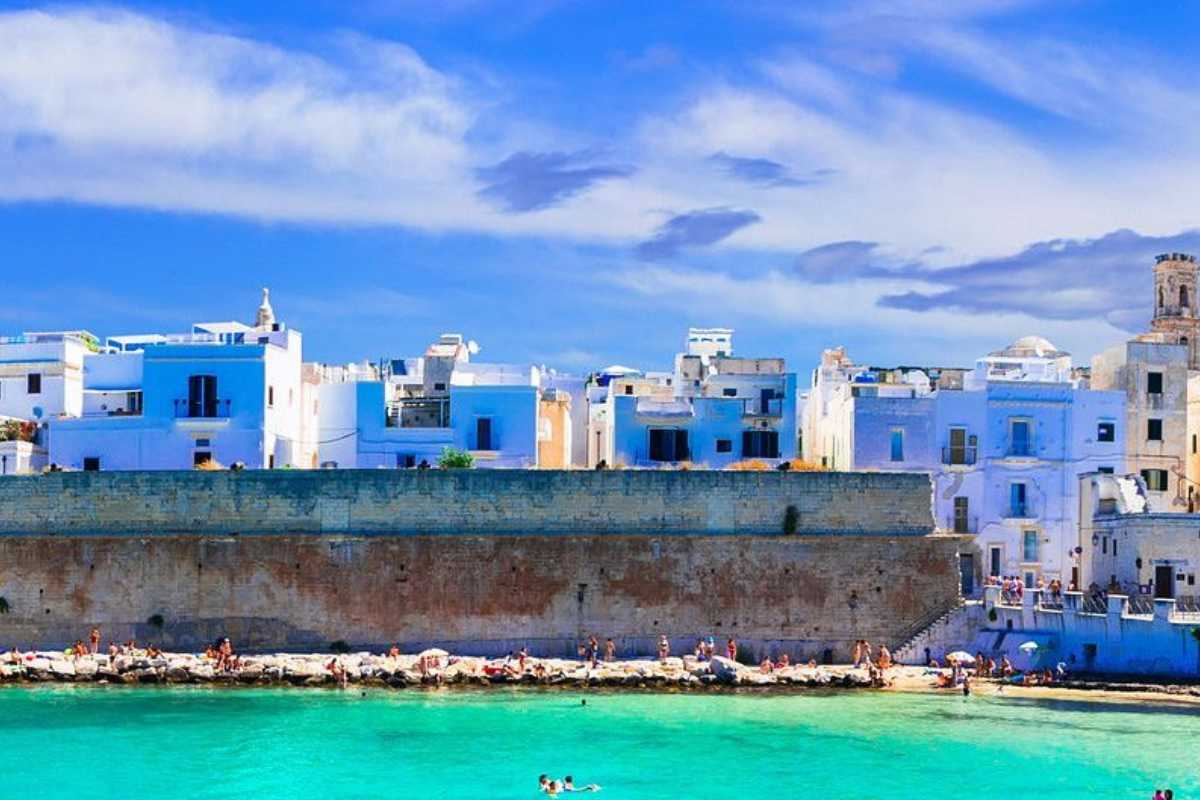 Dettaglio della stazione ferroviaria di Monopoli, treno regionale in arrivo, viaggiatori con valigie, atmosfera di viaggio estivo